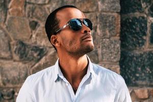 Man in style. Handsome young African man in sunglasses looking up while standing against the stoned wall outdoors photo