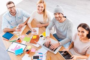 Creative team t work. Top view of group of business people in smart casual wear working together and smiling while sitting at the wooden desk photo