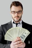 Confident and rich. Confident young man in formalwear and glasses holding paper currency and looking at camera while standing against grey background photo