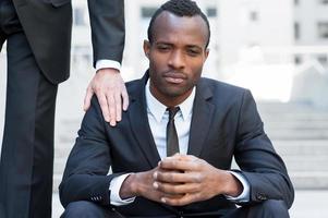 He needs a friendly support. Frustrated young African man in formalwear sitting on staircase while someone touching his shoulder with hand photo