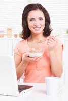 tener un desayuno saludable. hermosa joven tomando un desayuno saludable y sonriendo mientras se sienta en la mesa de la cocina con una laptop en ella foto