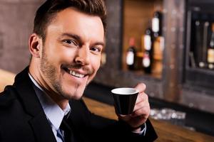Inspired by cup of fresh coffee. Confident young man in formalwear drinking coffee and smiling while sitting at the bar counter photo