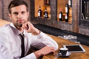 Surfing the net in bar. Handsome young man in shirt and tie holding hand on chin and smiling while sitting at the bar counter with digital tablet laying near him photo