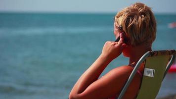 vrouw zittend Aan een dek stoel Aan de strand en pratend Aan cel telefoon video