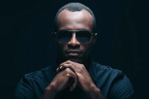 Confident and trendy look. Portrait of handsome young African man in sunglasses leaning his head on hands and looking at camera while being in front of black background photo