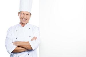 Cheerful chef. Confident mature chef in white uniform leaning at the copy space and smiling while standing against white background photo