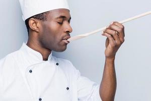 Chef trying meal. Side view of confident young African chef in white uniform keeping eyes closed while trying eating from wooden spoon and standing against grey background photo