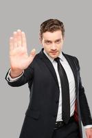 Stop it Serious young man in formalwear showing his palm and looking at camera while standing against grey background photo