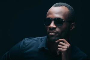 Cool and trendy. Portrait of handsome young African man in sunglasses holding hand on chin and looking away while being in front of black background photo