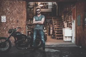 Welcome to my world Full length of confident young man keeping arms crossed and looking at camera while standing near his motorcycle garage or repair shop photo