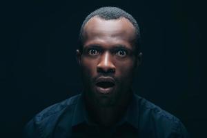 No way Portrait of surprised young African man looking at camera and keeping mouth open while being in front of black background photo