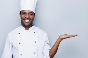 cocinero alegre. confiado joven chef africano en uniforme blanco sosteniendo espacio de copia y sonriendo mientras está de pie contra el fondo gris foto