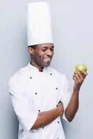 Cooking only healthy food. Confident young African chef in white uniform holding green apple and looking at it with smile while standing against grey background photo