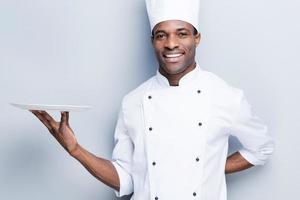 Copy space at his plate. Confident young African chef in white uniform holding empty plate and smiling while standing against grey background photo