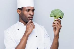 Choosing the best ingredient for his meal. Thoughtful young African chef in white uniform holding broccoli and looking at it while standing against grey background photo