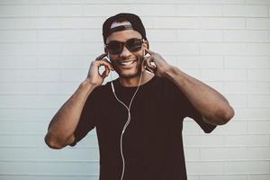 Music is my lifestyle Happy young African man adjusting his headphones and looking at camera while standing against the stoned wall outdoors photo