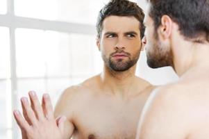 Is that acne Serious young shirtless man leaning at the mirror and looking at himself attentively photo