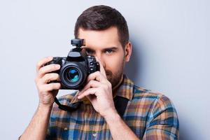 retrato sonriente de un joven confiado en camisa sosteniendo una cámara frente a su cara mientras se enfrenta a un fondo gris foto