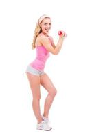 Me encantan los deportes de cuerpo entero de una hermosa mujer rubia pin-up con camisa rosa haciendo ejercicio con pesas y sonriendo mientras está de pie aislada en el fondo blanco foto