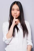 Be quiet Attractive young Asian woman looking at camera and holding finger on lips while standing against grey background photo