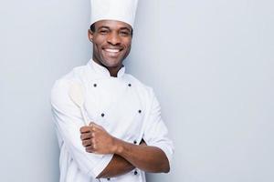 cocinero confiado. alegre joven chef africano con uniforme blanco que mantiene los brazos cruzados y sonríe mientras se enfrenta a un fondo gris foto