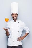 maestro culinario juguetón. confiado joven chef africano con uniforme blanco tirando pimienta y mirando a la cámara con una sonrisa mientras se enfrenta a un fondo gris foto