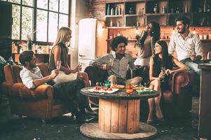 Friends will be friends. Group of cheerful young people enjoying food and drinks while spending nice time in cofortable chairs on the kitchen together photo