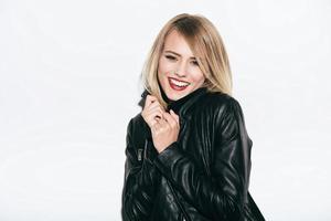 In her own style. Funky young woman in leather coat adjusting her collar and smiling while standing against white background photo