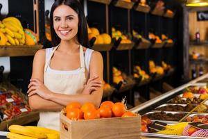 Welcome to fruit paradise Beautiful young woman in apron keeping arms crossed and smiling while standing in grocery store with variety of fruits in the background photo