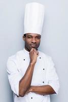 chef confiado y experimentado. Confiado joven chef africano en uniforme blanco sosteniendo la mano en la barbilla y mirando a la cámara mientras está de pie contra el fondo gris foto