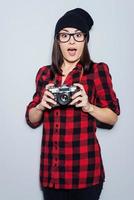 It can be a cool shot Surprised young woman in headwear and glasses holding camera and looking at camera while standing against grey background photo