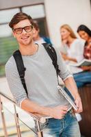 University life is great Smiling young man holding laptop and looking at camera while his friends standing in the background photo