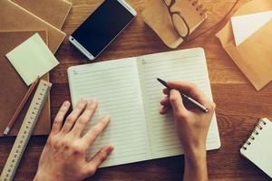 Just inspired. Close-up top view of man writing something in notebook while sitting at the wooden desk with different chancellery stuff laying around photo