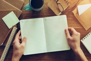 In search of inspiration. Close-up top view of male hands holding opened notebook over wooden desk with different chancellery stuff laying on it photo