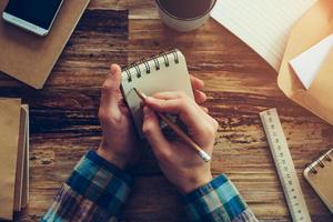 Making some urgent notes. Close-up top view of man holding notebook and writing something while sitting at wooden desk with different chancellery stuff laying on it photo