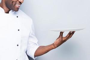 Chef with plate. Close-up of confident young African chef in white uniform holding empty plate and smiling while standing against grey background photo