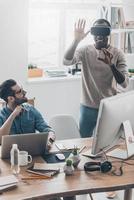 Enjoying new reality Two young successful men in casual wear working in creative office while one of them standing in VR headset photo
