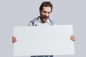 Wow Young confident man in smart casual clothes holding blank flipchart with surprised face while standing against grey background photo