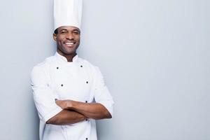 cocinar es mi pasión. confiado joven chef africano en uniforme blanco con los brazos cruzados y sonriendo mientras está de pie contra el fondo gris foto