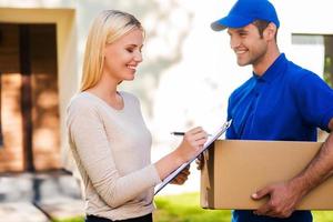 Sign here please Smiling young delivery man holding a cardboard box while beautiful young woman putting signature in clipboard photo