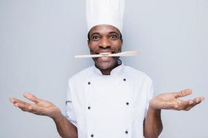 Surprised chef. Surprised young African chef in white uniform holding wooden spoon in his mouth while standing against grey background photo