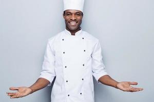 Welcome in the world of tastes. Confident young African chef in white uniform keeping arms outstretched and smiling while standing against grey background photo