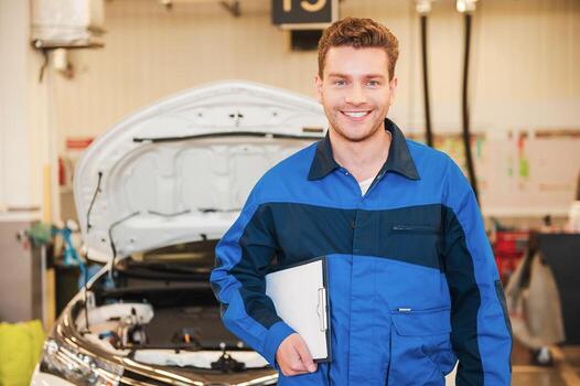 How may I help you Handsome young man in uniform holding clipboard and smiling while standing in workshop with car in the background photo