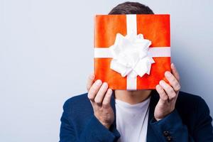 Gift man. Close-up of young man in formalwear covering his face by a gift while standing against grey background photo