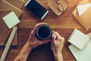 Coffee break. Close-up top view of male hands holding cup with coffee while sitting at the wooden desk with different chancellery laying around photo