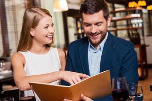 Try this meal Beautiful young loving couple examining menu and smiling while sitting at the restaurant together photo