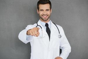 cuidaré de su salud, un joven y alegre doctor con uniforme blanco que apunta a la cámara mientras se enfrenta a un fondo gris foto