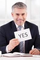 Approved Cheerful grey hair man in formalwear holding a paper with yes sign on it while sitting at her working place photo