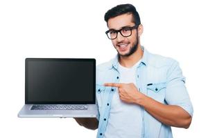 This is what you need Confident young Indian man carrying laptop on hand and pointing it while standing against white background photo