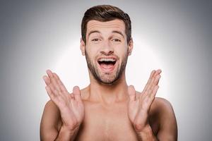 Estoy tan feliz retrato de un joven feliz sin camisa mirando a la cámara y gesticulando mientras está de pie contra un fondo gris foto